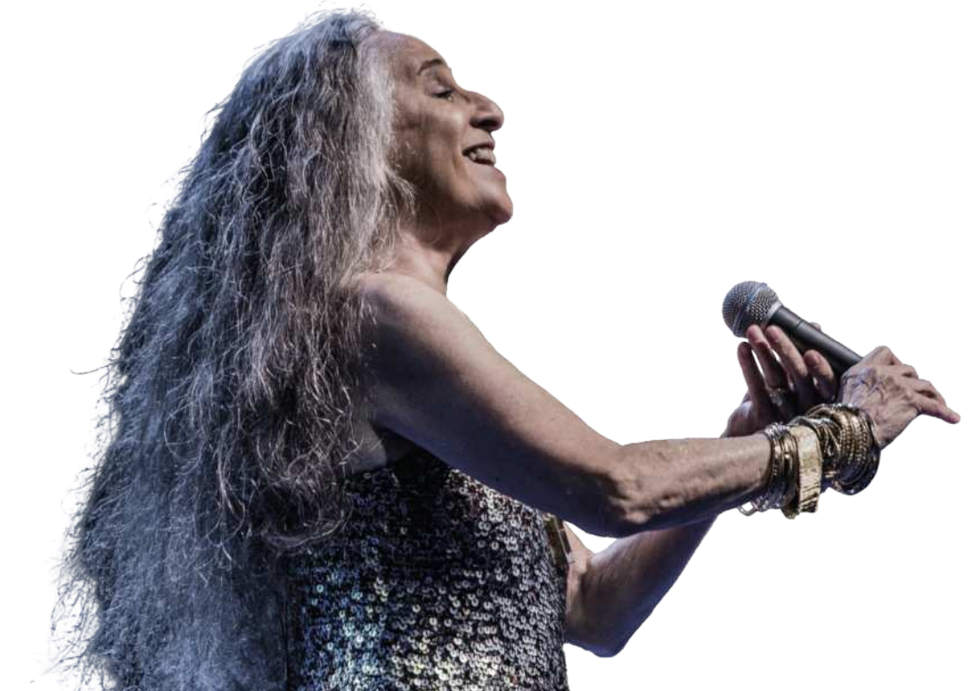Foto de Maria Bethânia sorrindo e segurando um microfone. Ela possui cabelos compridos, ondulados e grisalhos.