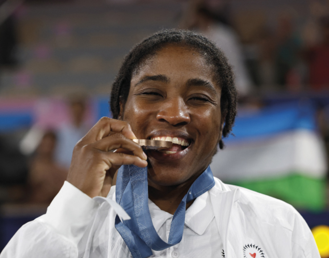 Foto de Cindy Ngamba. Ela é uma jovem negra e comemora mordendo a medalha de bronze que recebeu.