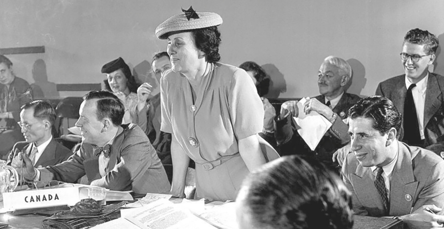 Foto de homens e mulheres reunidos em um salão, representando diferentes países. Dentre eles, destaca-se Bertha Lutz em pé, uma mulher branca usando vestido, cinto e chapéu.