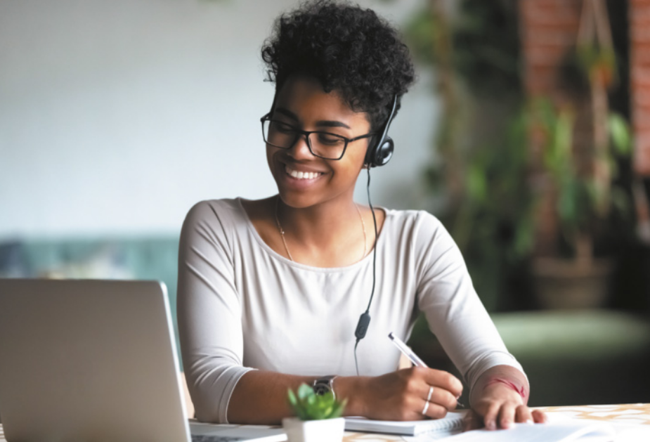 Imagem de mulher jovem escrevendo em um caderno. Ela usa fones de ouvido e está diante de um computador.