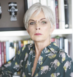 Fotografia de Lívia Barbosa. Ela é branca, possui cabelos brancos curtos, usa óculos e camisa estampada, está com o cotovelo direito apoiado em uma estante com livros.