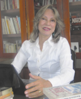 Fotografia de Elisabeth da Fonseca Guimarães sorrindo. Ela é branca, loira, usa camisa de mangas compridas e está sentada diante de um notebook.
