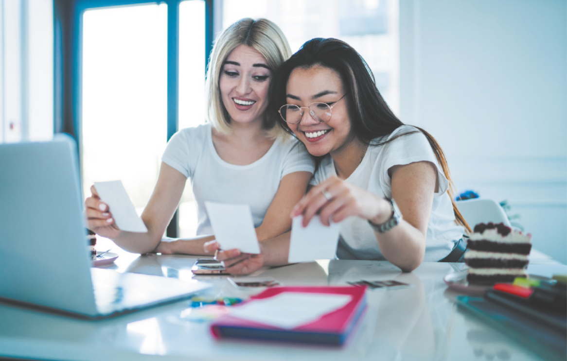 Imagem de duas jovens sentadas diante de uma mesa, segurando cartões de papel. Sobre a mesa, há, um computador, um caderno, um celular e uma fatia de bolo.
