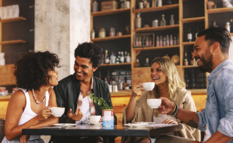 Imagem de dois homens e duas mulheres. Eles estão sentados ao redor de uma mesa, tomando café servido em xícaras.