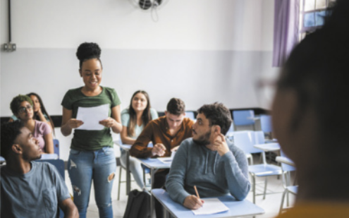 Imagem de sala de aula. Os estudantes estão sentados diante de carteiras, organizadas em fileiras. Entre elas, uma jovem está em pé, lendo uma folha de papel.