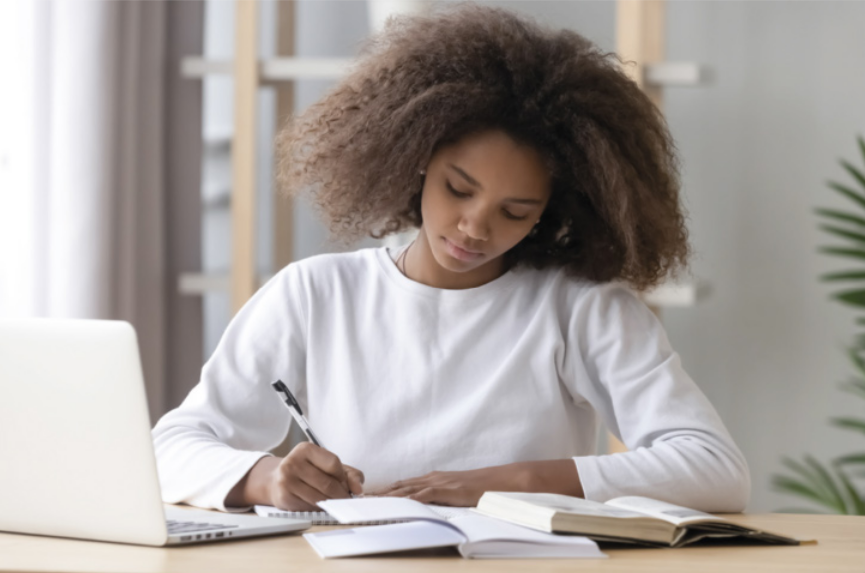 Imagem de uma jovem fazendo anotações em um caderno. Diante dela, há um notebook e dois livros abertos.