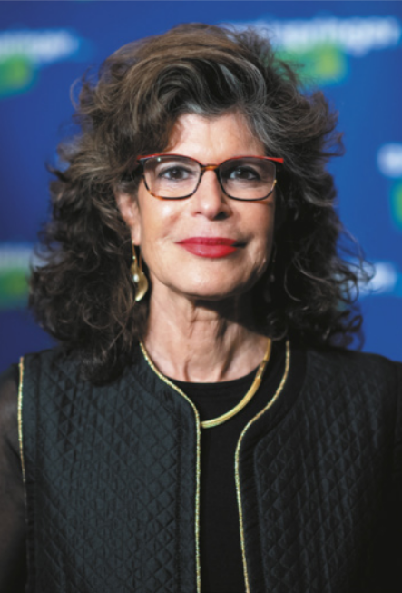 Fotografia de Shoshana Zuboff. Ela é branca, possui cabelos ondulados que se estendem até os ombros e usa óculos, brincos, batom e colete.