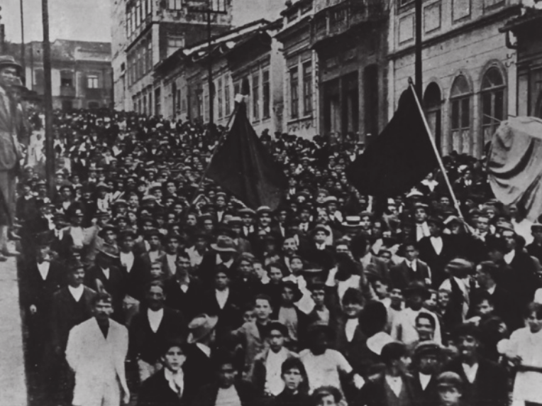 Imagem em preto e branco de multidão descendo uma ladeira. Duas pessoas carregam bandeiras.