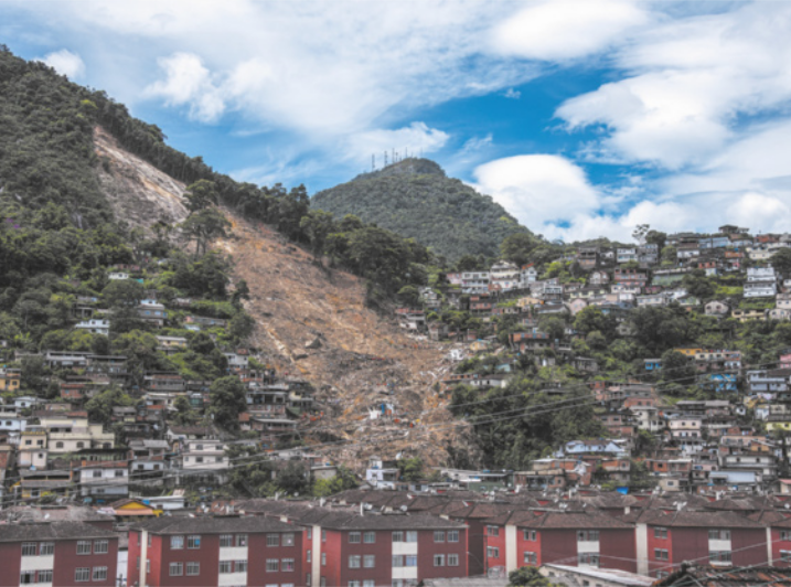 Imagem de casas na encosta de um morro. Parte do terreno desabou, acumulando escombros no sopé do morro.