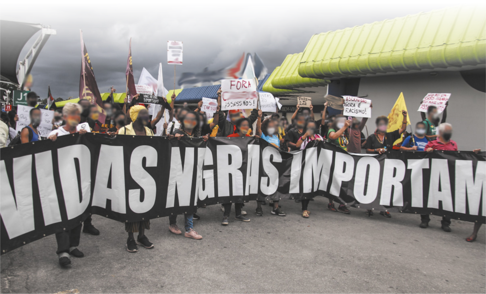 Imagem de manifestantes caminhando em uma rua com bandeiras e cartazes. As pessoas à frente do grupo carregam uma faixa com a seguinte frase: vidas negras importam.
