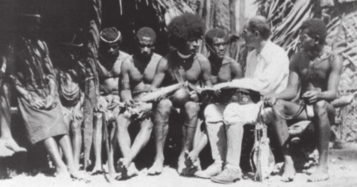 Fotografia em preto e branco de Bronislaw Malinowski com um grupo de homens indígenas. Eles estão sentados lado a lado em um banco e Malinowski está com maleta sobre as pernas.