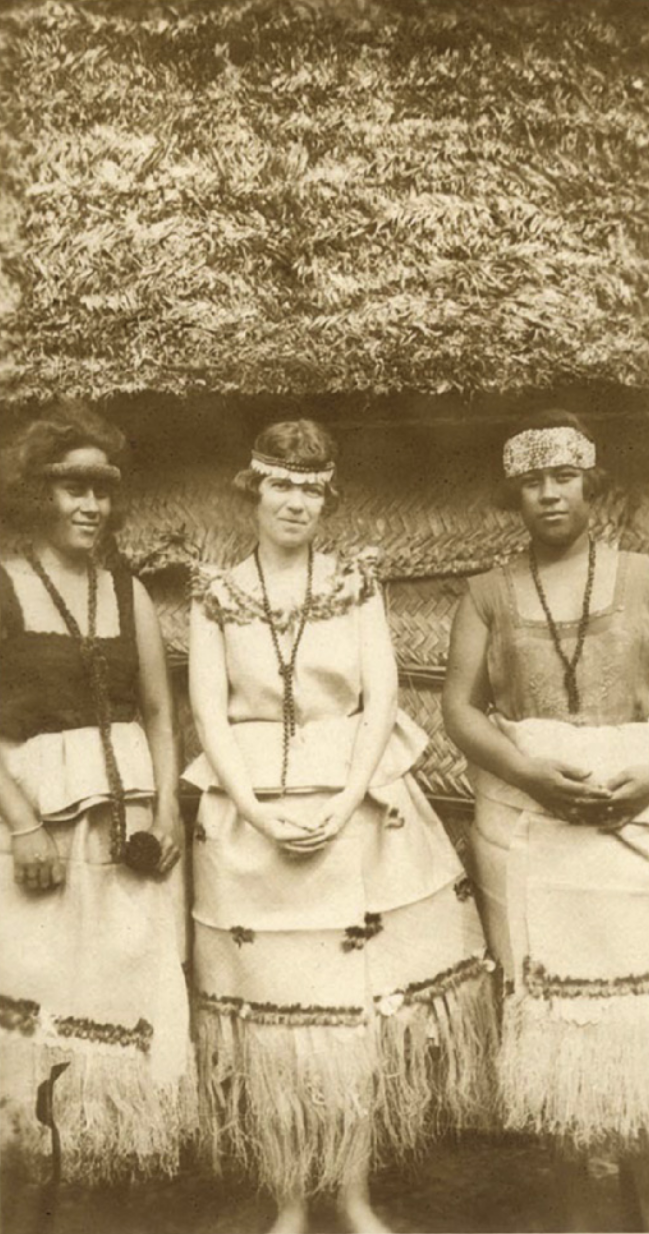 Fotografia em preto e branco de Margaret Mead entre duas jovens indígenas. Elas usam faixa ao redor da cabeça, colar de contas, regata e saia com a barra feita de palha.