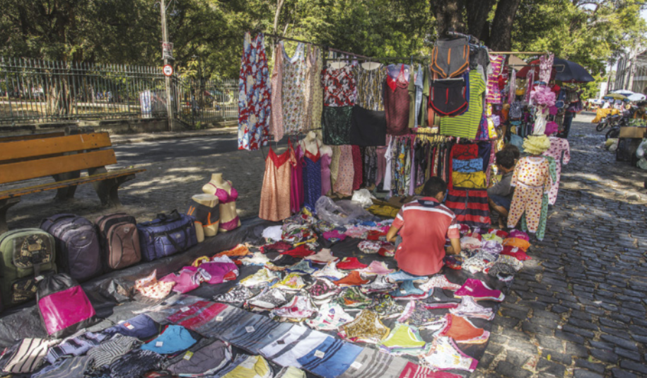 Imagem de roupas, bolsas e mochilas à venda em uma praça. Os produtos estão expostos em barracas e em uma lona estendida no chão.