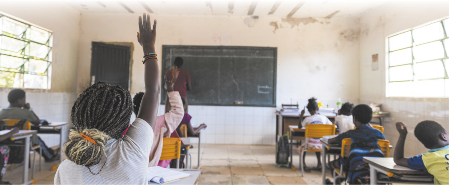 Imagem de professor e estudantes negros em uma sala de aula. O professor faz anotações na lousa e os estudantes estão sentados diante de carteiras, organizadas em fileiras. Um estudante está com o braço direito erguido.