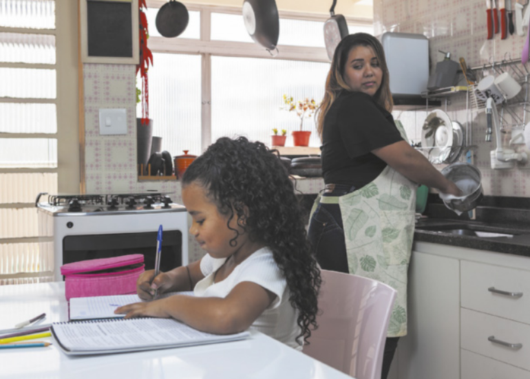 Imagem de mãe e filha em uma cozinha. A filha está sentada diante da mesa, escrevendo em um caderno. A mãe está lavando louça, com o rosto voltado para trás, em direção à filha.