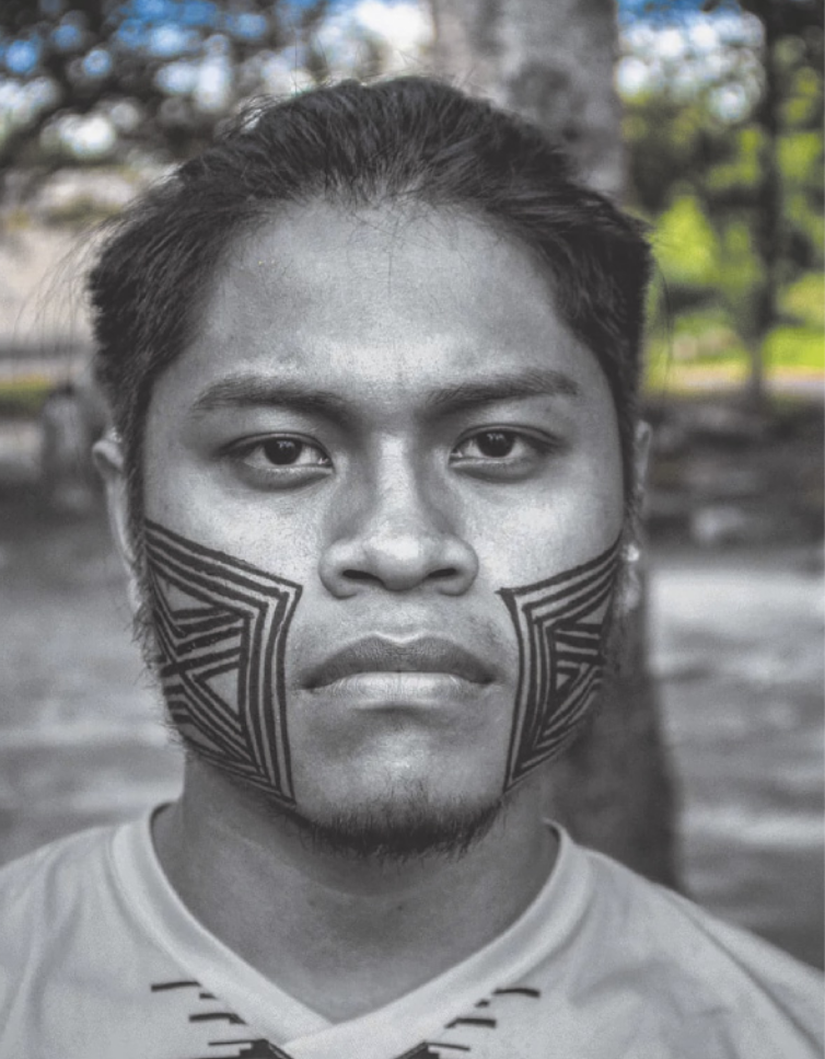 Fotografia em preto e branco de Cristian Wariu. Ele é indígena, possui cabelo curto e está com uma sequência de quadrados pintada em cada bochecha. No centro dos quadrados, há linhas perpendiculares que formam um X.