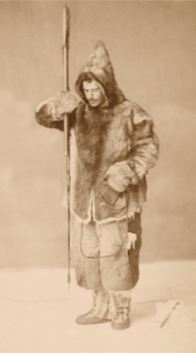Fotografia em preto e branco de Franz Boas. Ele segura uma lança com a ponta voltada para baixo e usa casaco de pele com capuz, luvas, calça ampla e botas.