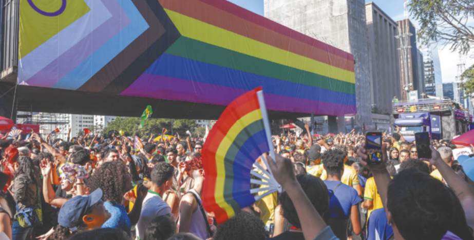 Imagem de evento público coletivo em uma avenida. As pessoas seguram bandeirinhas e objetos com as cores do arco-íris.