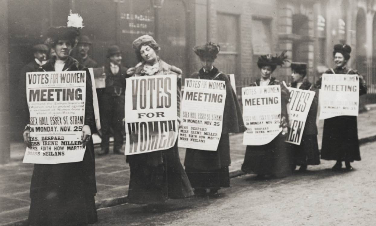 Fotografia em preto e branco de sufragistas. Elas caminham enfileiradas em uma rua, carregando cartazes com frases em inglês.