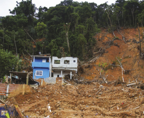 Imagem de terra, escombros e troncos de árvores na encosta de um morro. À esquerda, há um sobrado parcialmente destruído.