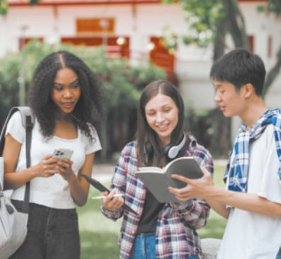 Imagem de três estudantes ao ar livre. Um deles mostra um livro para as colegas, que utilizam tablet e celular.
