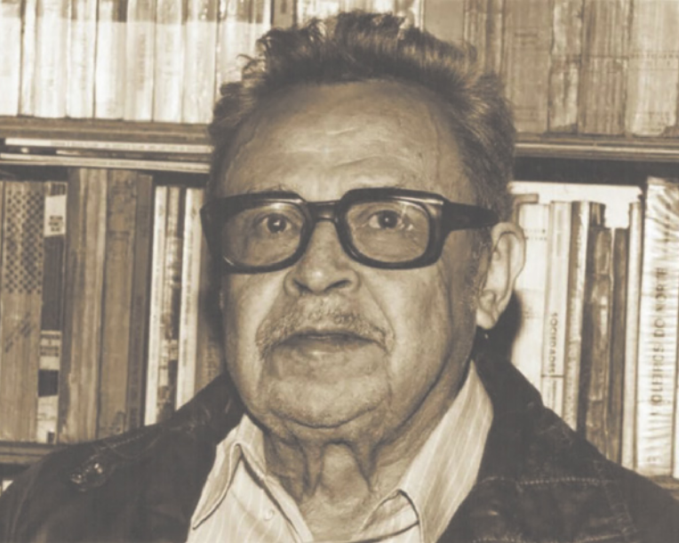 Fotografia em preto e branco de Clovis Moura. Ele é negro, idoso, possui cabelo curto e bigode, usa óculos e está de costas para uma estante com livros.