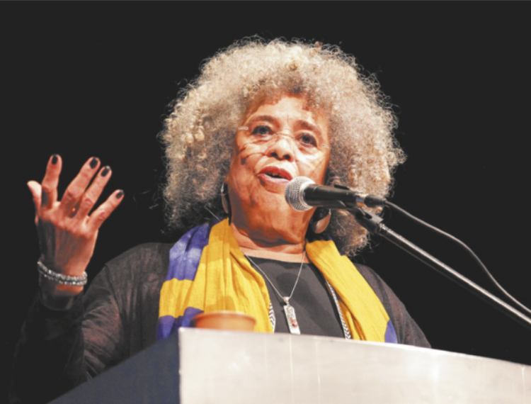 Imagem de Angela Davis. Ela é negra, idosa, usa óculos, está em um púlpito com microfone e gesticula com a mão direita.