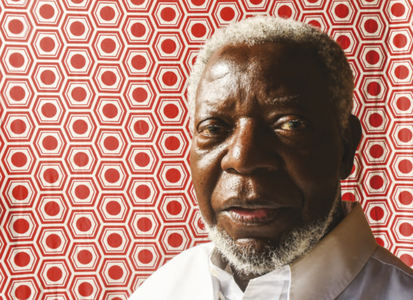 Fotografia de Kabengele Munanga. Ele é negro, idoso, possui barba e está diante de um painel com estampa geométrica.