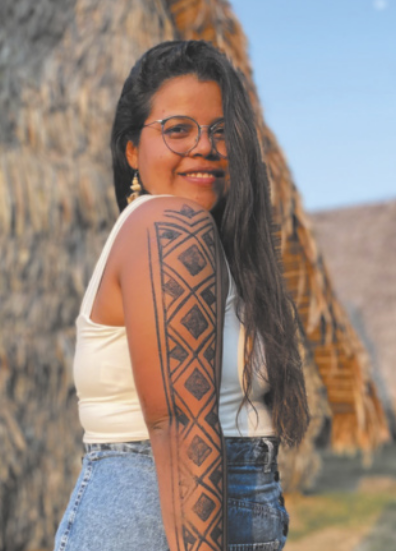 Fotografia de Odenilze Ramos sorrindo. Ela é jovem, possui cabelos longos, usa óculos e está com pintura geométrica no braço esquerdo.