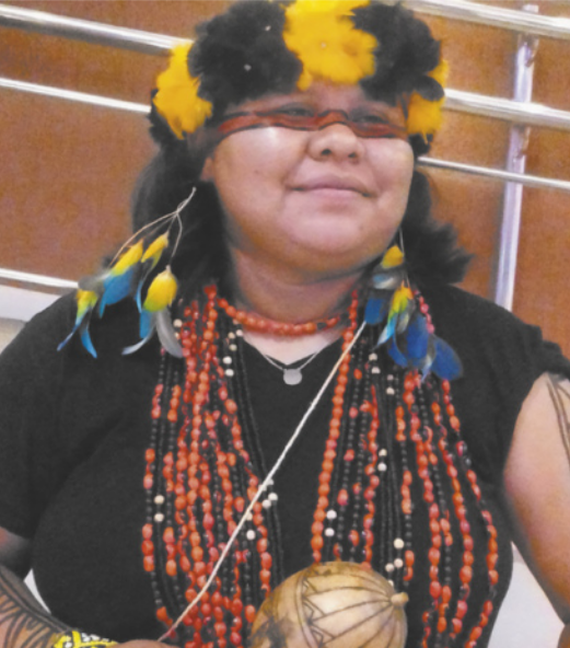 Fotografia de Tipuici Manoki sorrindo. Ela é indígena, usa coroa e brincos de penas coloridas, colares de contas e camiseta, está com pintura corporal e segura um chocalho.