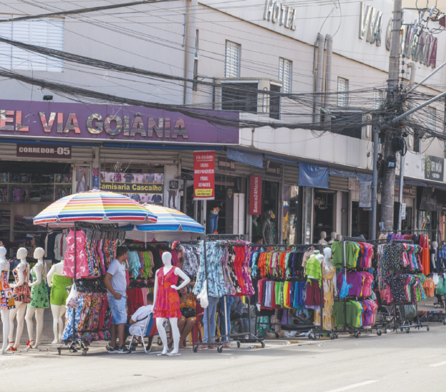 Imagem de rua comercial. Roupas estão penduradas em cabideiros ao longo da calçada.