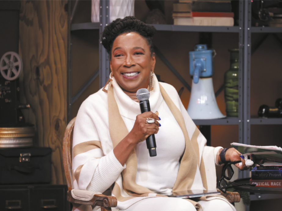 Fotografia de Kimberlé Crenshaw sorrindo. Ela é negra, possui cabelo curto, está sentada em uma cadeira, segura um óculos e utiliza um microfone.