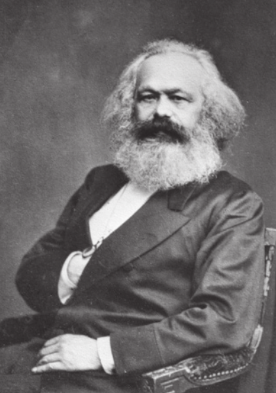 Fotografia em preto e branco de Karl Marx. Ele pé branco, possui cabelo, barba e bigode grisalhos e abundantes, está sentado em uma cadeira e apoia a mão direita da barriga, com os dedos dentro do paletó.