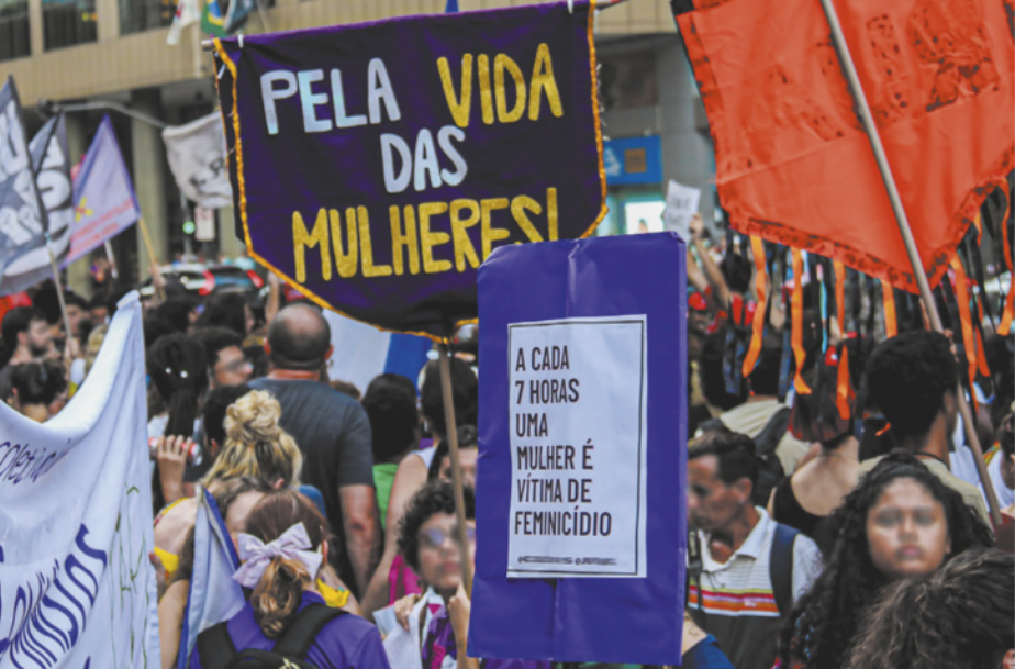 Imagem de manifestantes com cartazes e estandartes. Um dos cartazes contém a seguinte frase: a cada 7 horas uma mulher é vítima de feminicídio. Um dos estandartes contém a seguinte frase: pela vida das mulheres!