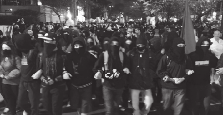 Fotografia em preto e branco de manifestantes. Eles usam boné, gorro ou capuz, estão com o rosto coberto e caminham de braços dados.