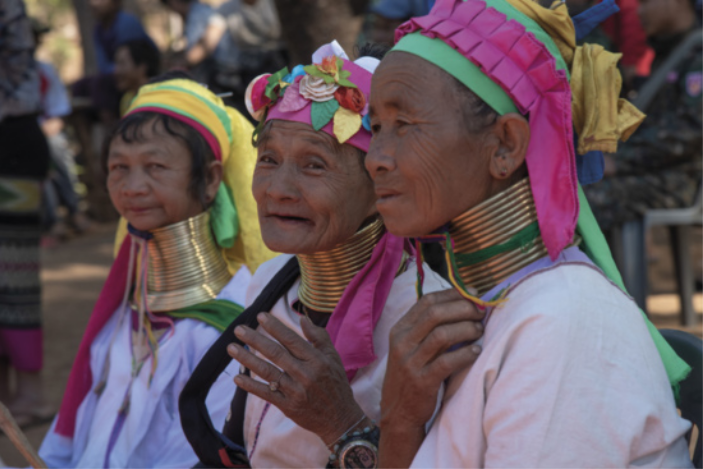 Imagem de três mulheres indígenas. Elas usam faixas coloridas ao redor da cabeça e diversas argolas sobrepostas, que alongam o pescoço.