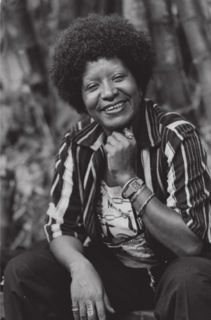 Fotografia em preto e branco de Lélia Gonzalez sorrindo. Ela é negra, possui cabelos crespos, usa camisa listrada e está sentada, com o queixo apoiado sobre a mão esquerda.