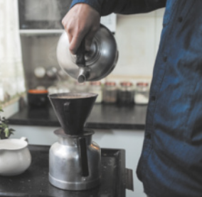 Imagem de pessoa coando café. Ela despeja a água quente em um coador, que está encaixado em um bule.