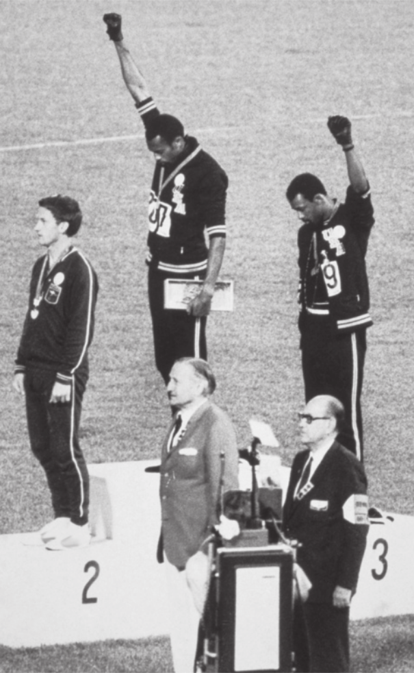 Fotografia em preto e branco de atletas em um pódio. O segundo lugar é ocupado por um atleta branco. O primeiro e o terceiro lugares são ocupados por atletas negros, que estão com o braço erguido e o punho fechado. Em frente ao pódio, há dois homens brancos com terno e gravata.