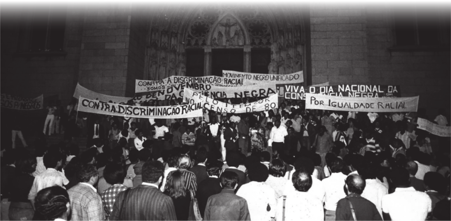 Imagem de manifestantes em frente a uma catedral. Eles carregam faixas com as seguintes inscrições: contra a discriminação racial; por igualdade racial; viva o Dia Nacional da Consciência Negra; Movimento Negro Unificado.
