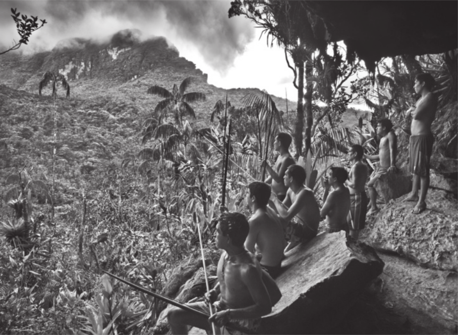 Fotografia em preto e branco de homens indígenas armados com lanças. Eles estão apoiados em pedras na encosta de uma montanha com cobertura vegetal densa.