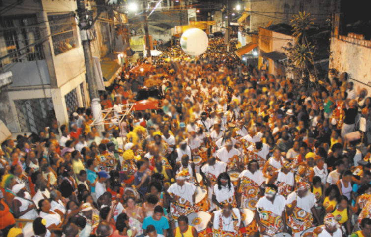 Imagem de multidão em uma rua. Ao centro, um grupo de pessoas toca tambores.