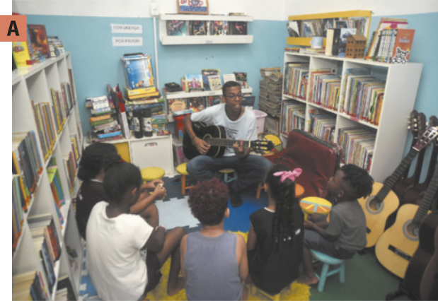 A. Imagem de professor tocando violão para os estudantes. Eles estão sentados em banquinhos dispostos em semicírculos. Ao redor deles, há violóes apoiados nas paredes, prateleiras e estantes com livros.