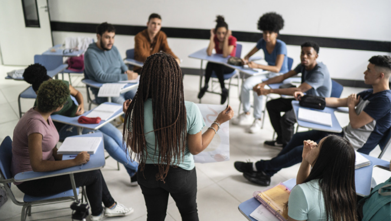Imagem de estudantes em uma sala de aula. Eles estão sentados em carteiras, dispostas em círculo. Uma estudante está em pé à frente do círculo e mostra um cartaz para os colegas.