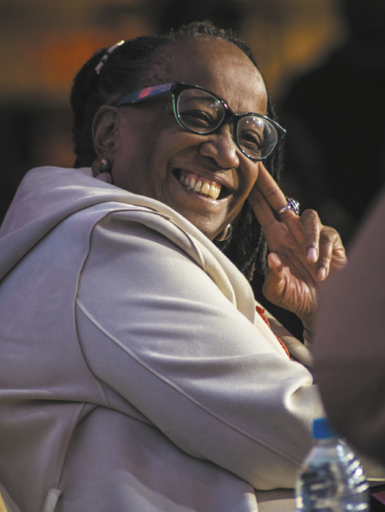 Fotografia de Sueli Carneiro sorrindo. Ela é negra, idosa, possui tranças nos cabelos e usa óculos.