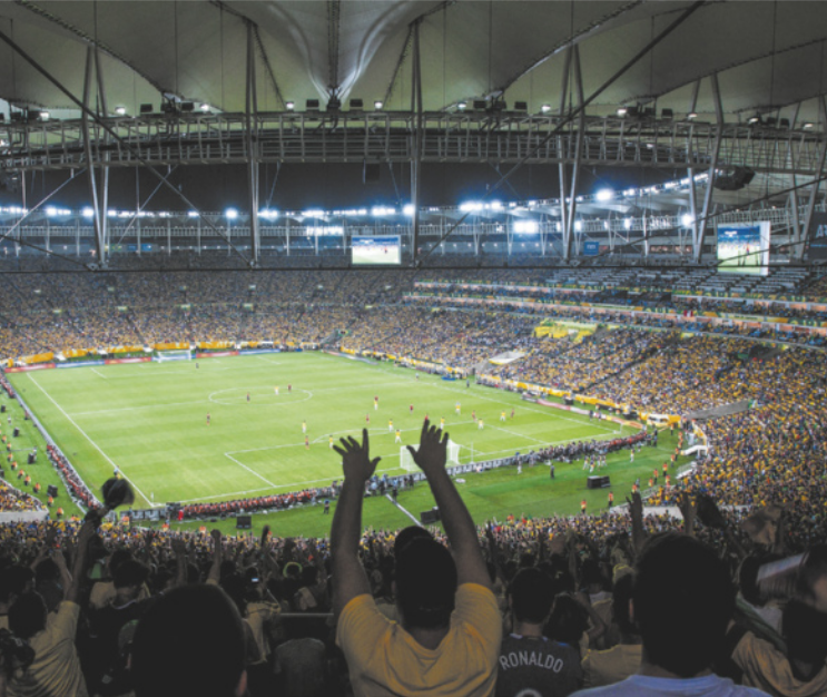 Imagem de jogo de futebol em um estádio. As arquibancadas estão lotadas e os torcedores erguem os braços.