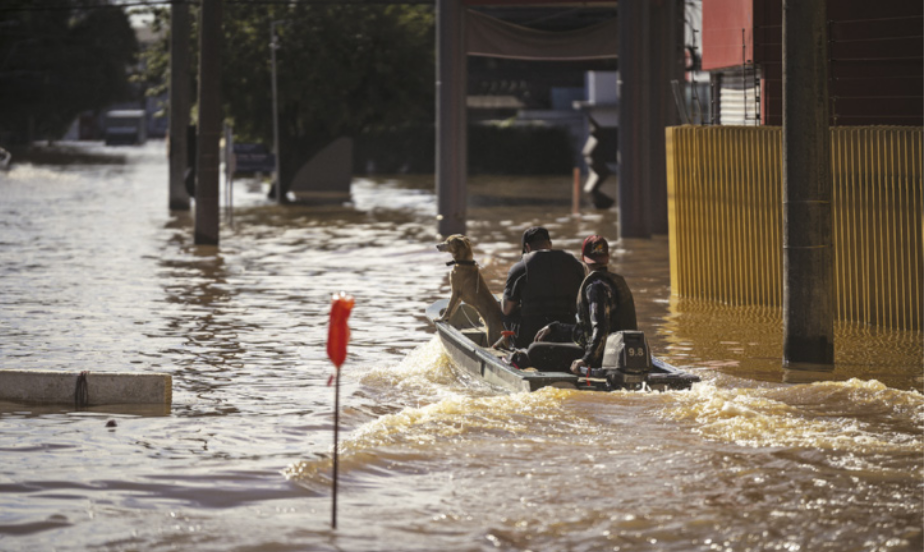 Imagem de área urbana alagada. Duas pessoas e um cachorro navegam em um barco motorizado.