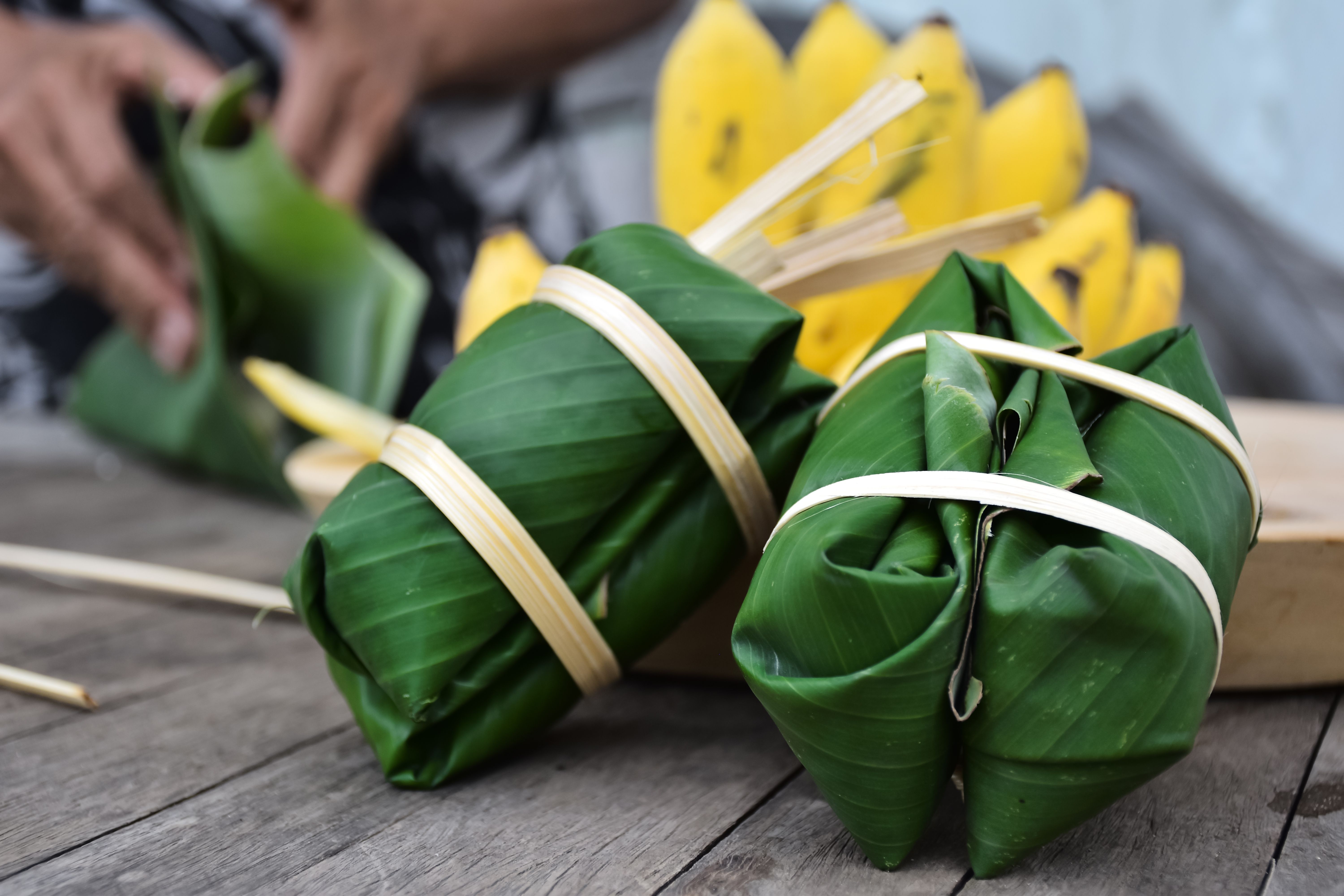 Fotografia. Dois pacotes de folhas de bananeiras verdes com fitas ao redor. Ao fundo, desfocado, há um cacho de bananas e as mãos de uma pessoa dobrando uma folha de bananeira.