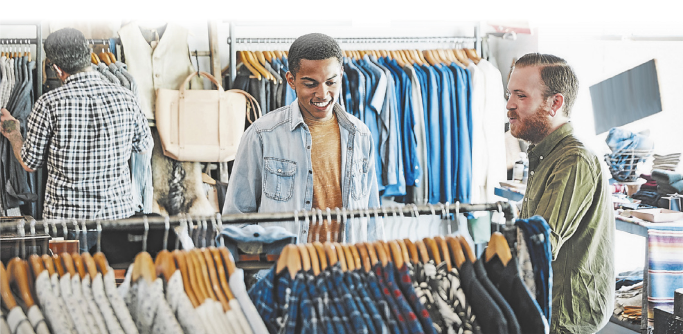 Fotografia de dois homens em uma loja de roupas. Eles estão sorrindo, e há uma arara com muitas roupas dispostas em cabides na frente de ambos. Ao fundo, mais araras de roupas e um homem olhando os produtos.