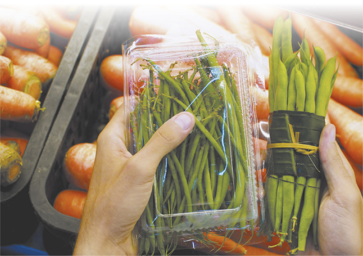 Fotografia das mãos de uma pessoa segurando alimentos. Com uma das mãos, a pessoa segura uma embalagem plástica contendo os alimentos. Com a outra mão, ela segura os alimentos amarrados com uma folha de bananeira. Ao fundo há caixotes com legumes.